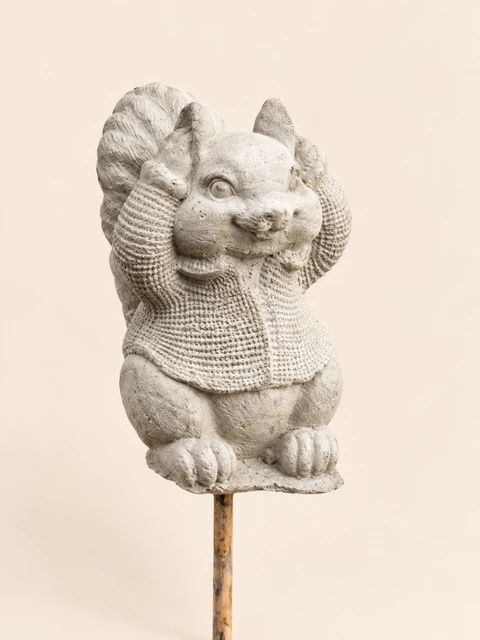 Light squirrel in a patterned sweater with its hands at its ears, on a slender stake