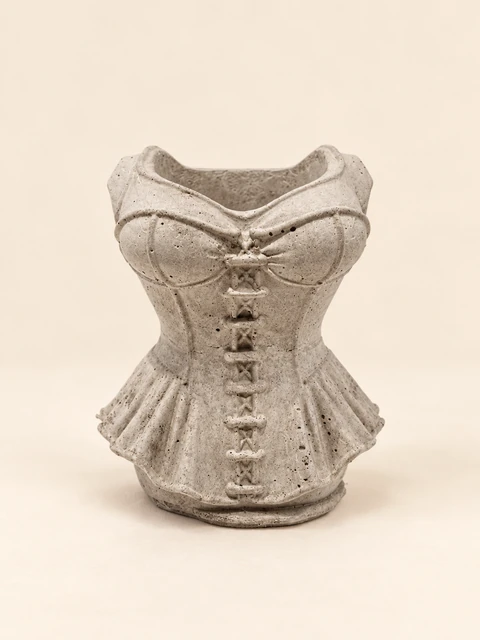 Corset container in natural concrete colour, with an open top, raised fastenings and folds at the lower edge